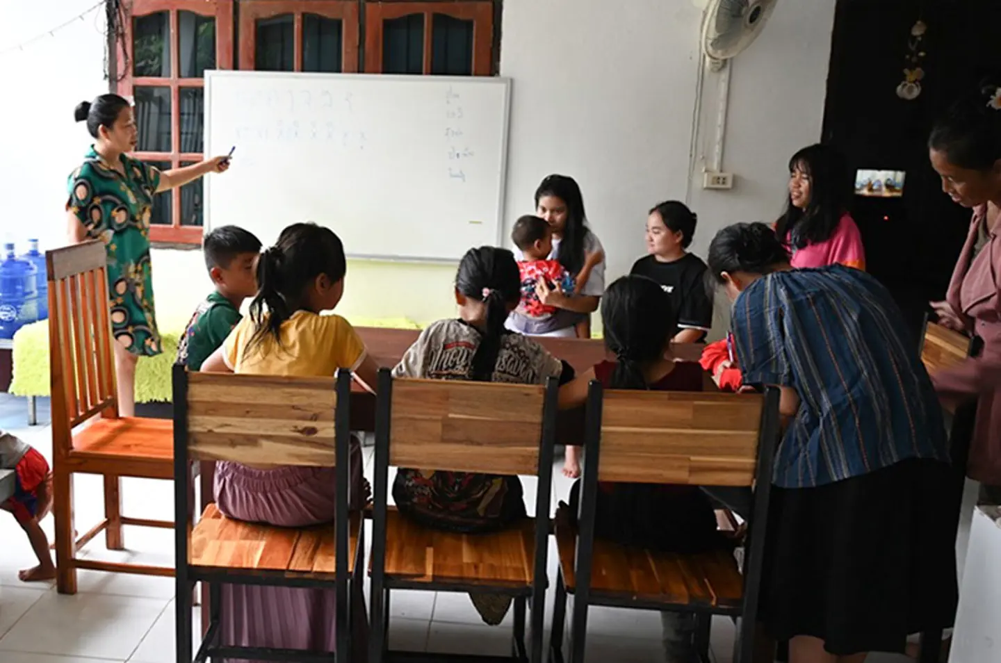 Adult woman teaching at a whiteboard while several girls and caregivers sit together in a classroom at a family home in Laos.